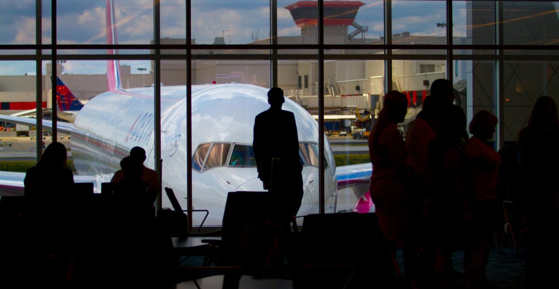Silhouettes of travelers waiting at an airport terminal with an airplane visible through the window.